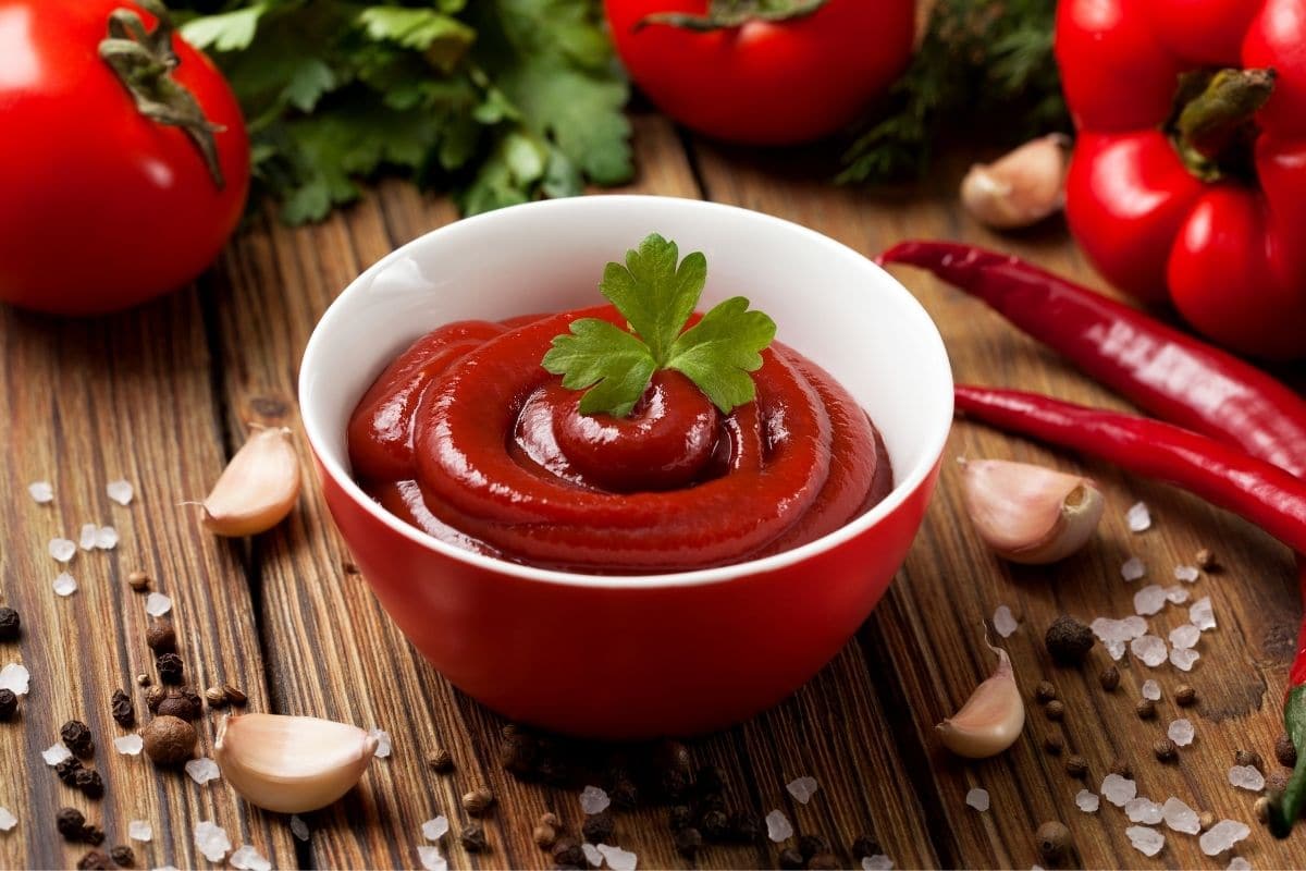 Red-white bowl of ketchup with hero on the top on table with ingredients scattered around.