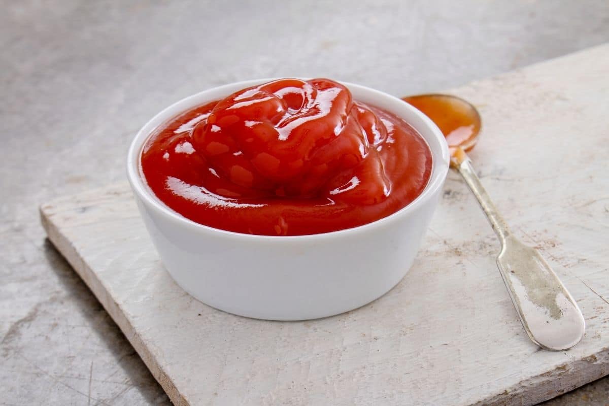 White bowl full of ketchup on white wooden board with spoon.
