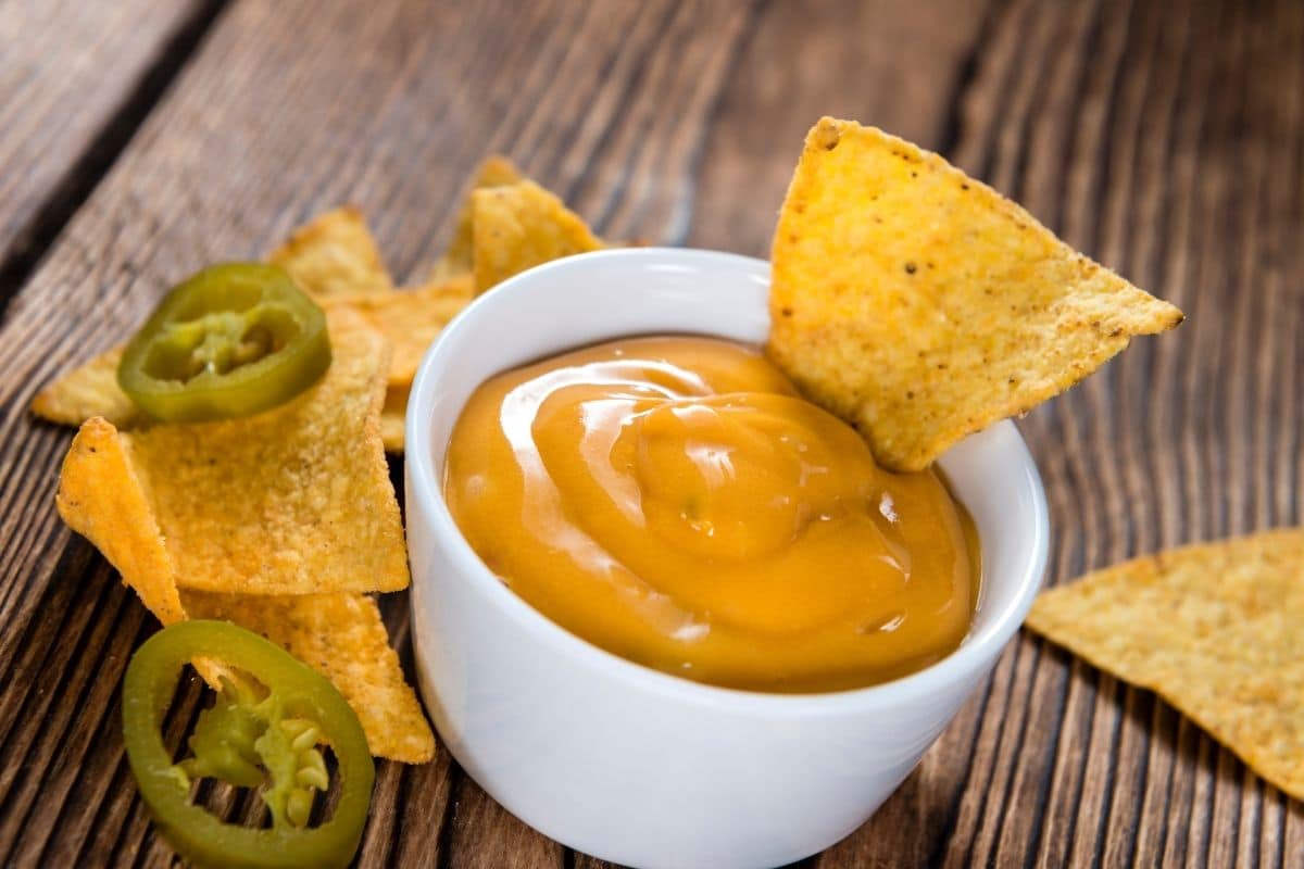 White bowl of nacho cheese on table with nachos and peppers.