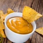 White bowl of nacho cheese with nacho on wooden table.