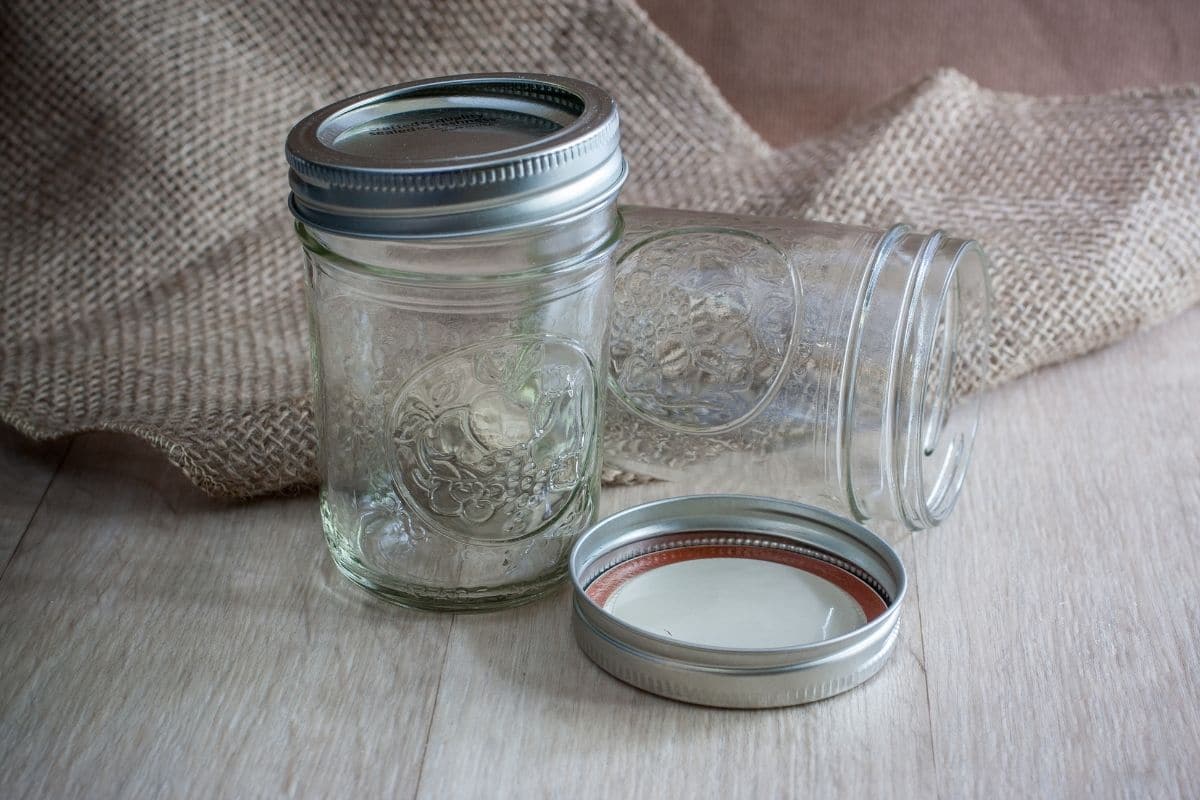 Two mason jars with lids on table with burlap in the background.