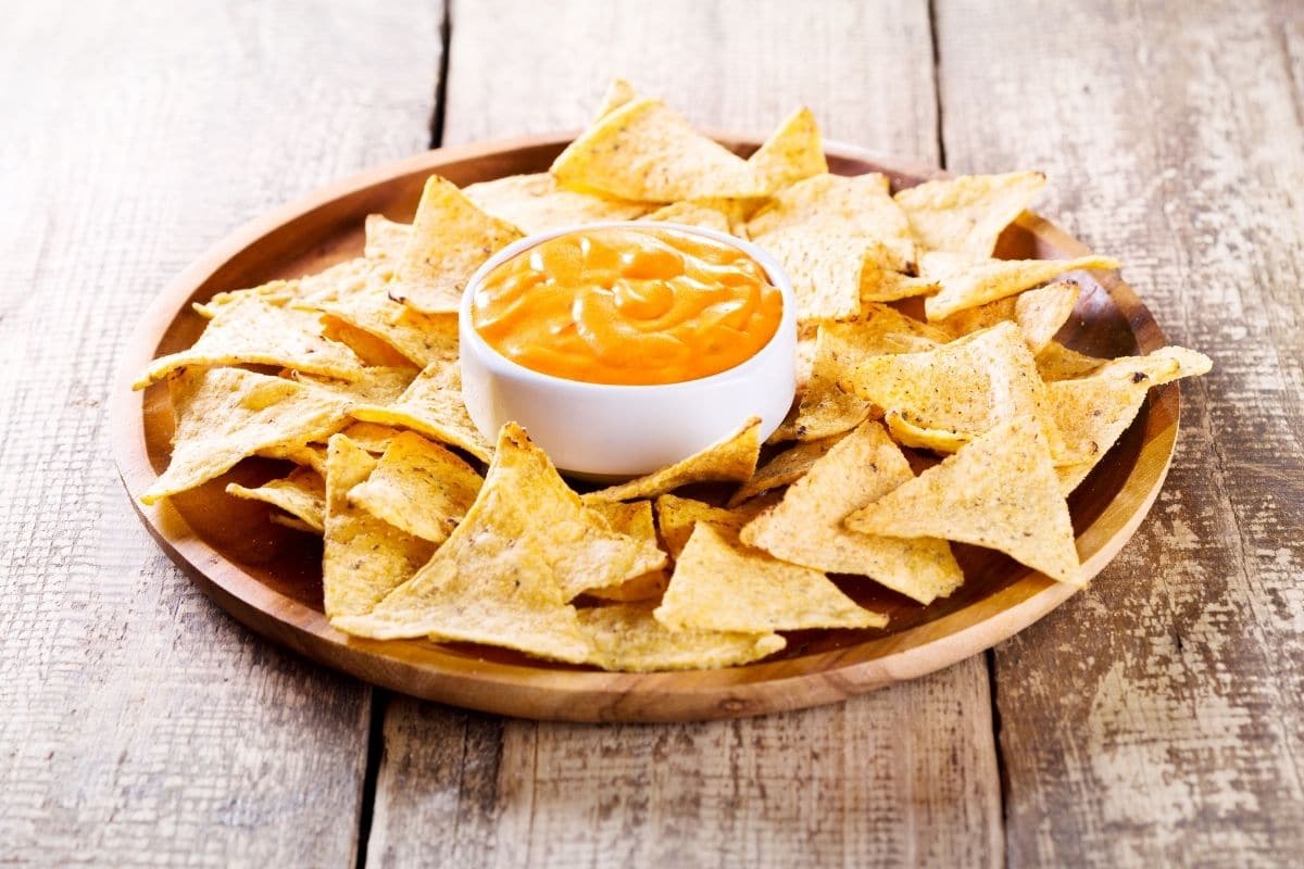 Tray full of nachos and bowl of nacho cheese on wooden table.