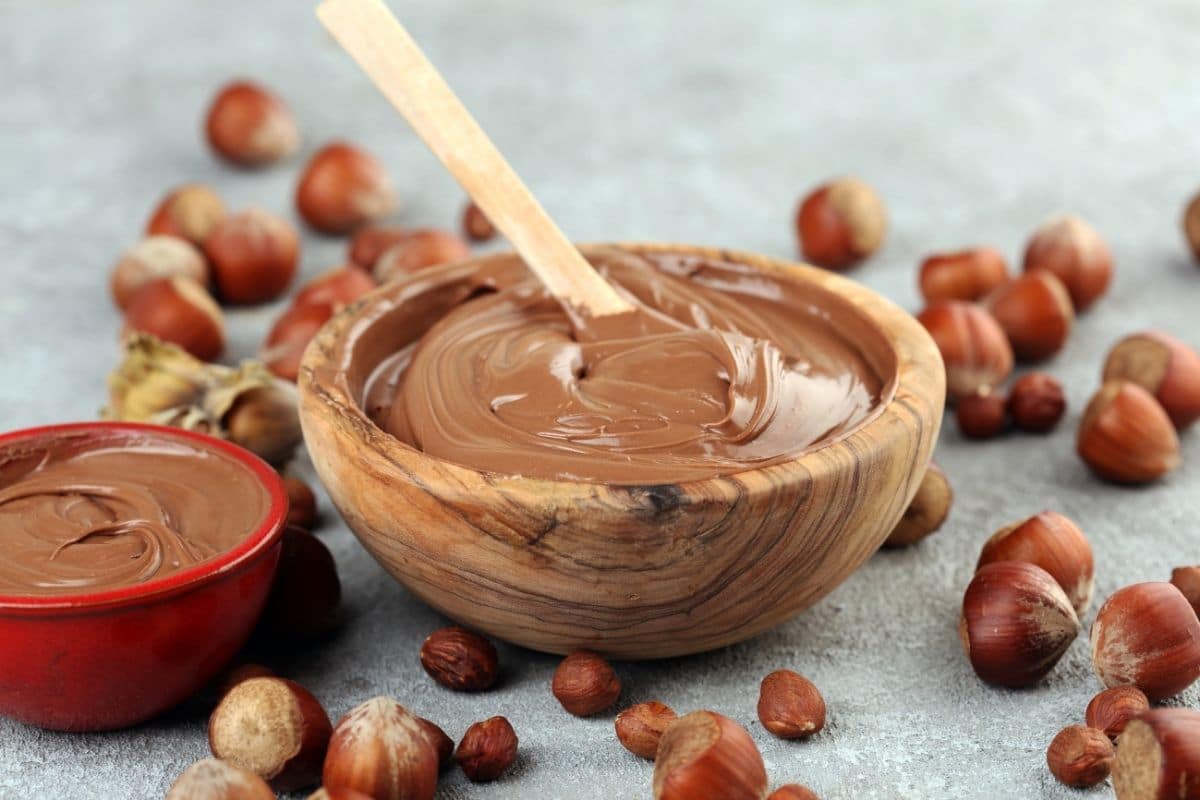 Wooden bowl of nutella with spoon on table with scattered hazelnuts around.