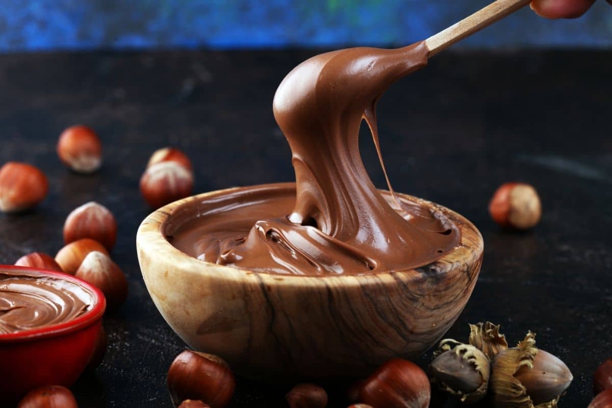 Wooden bowl of nutella with spoon over on black table with scattered hazelnuts around.