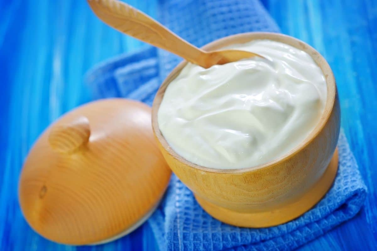 Wooden bowl of sour cream with spoon and lid on blue napkin.