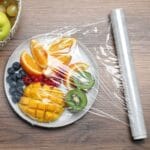 White plate full of fruit wraped by plastic wrap on brown table.