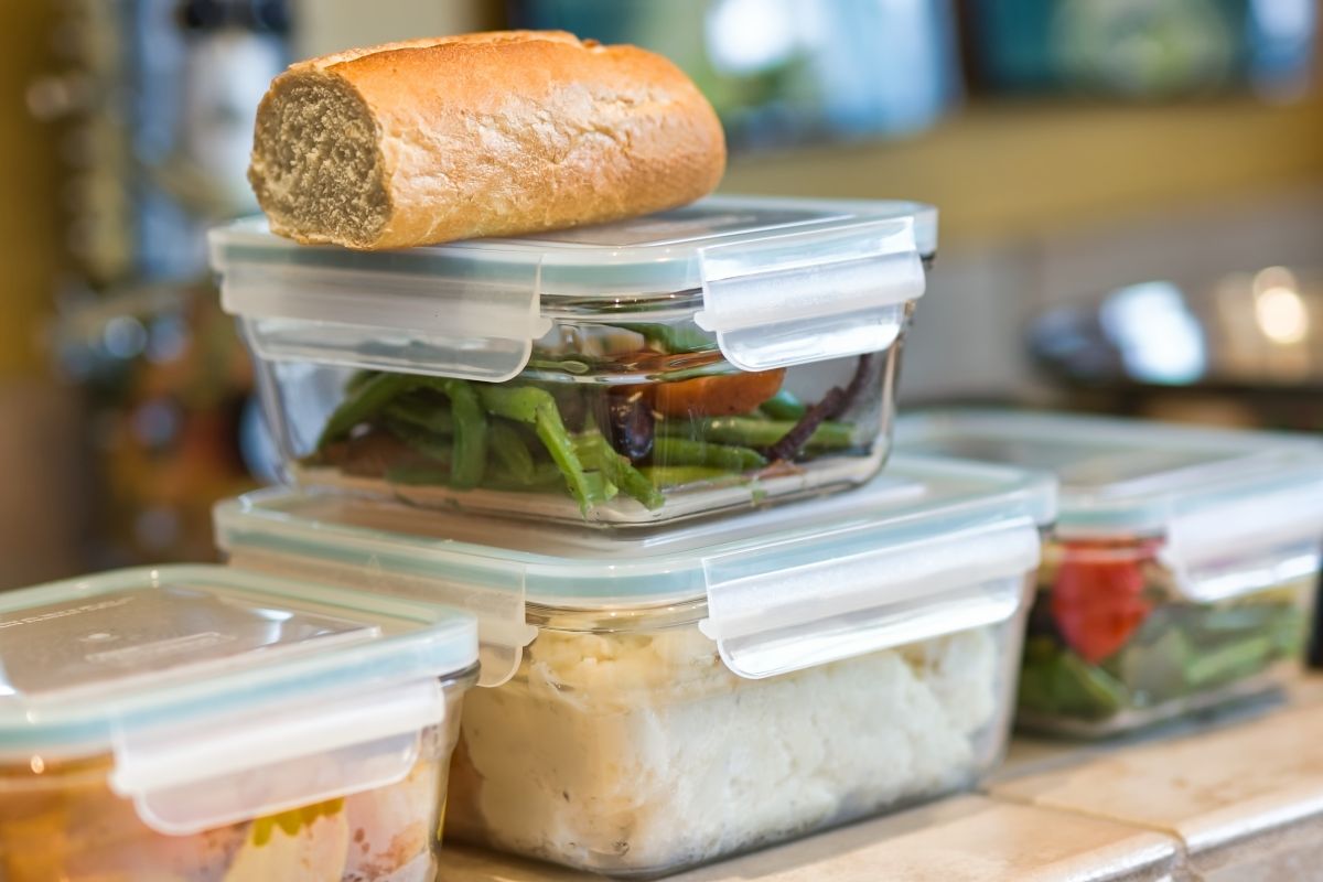 Bunch of plastic contianers with leftovers , loaf of bread on the top of them.