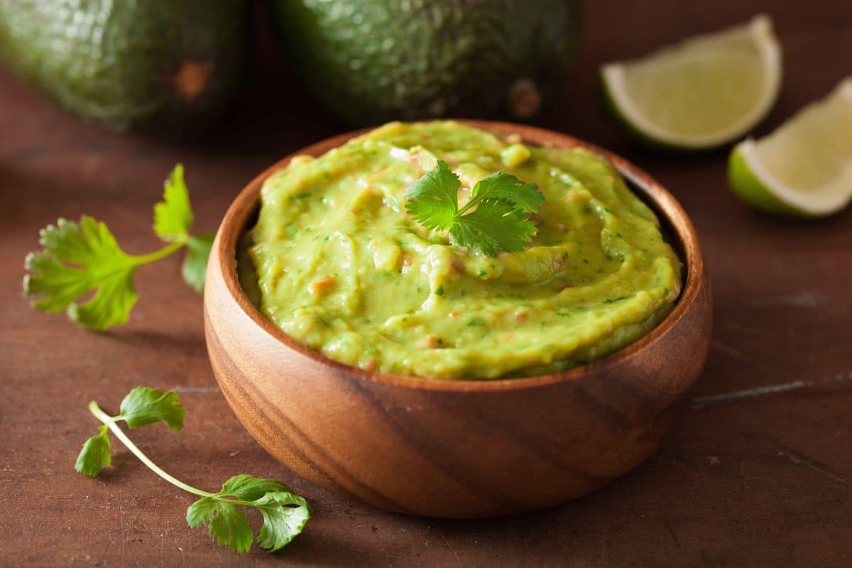 Wooden bowl of guacamole on brown table with herb, slices of lemon and avocados.
