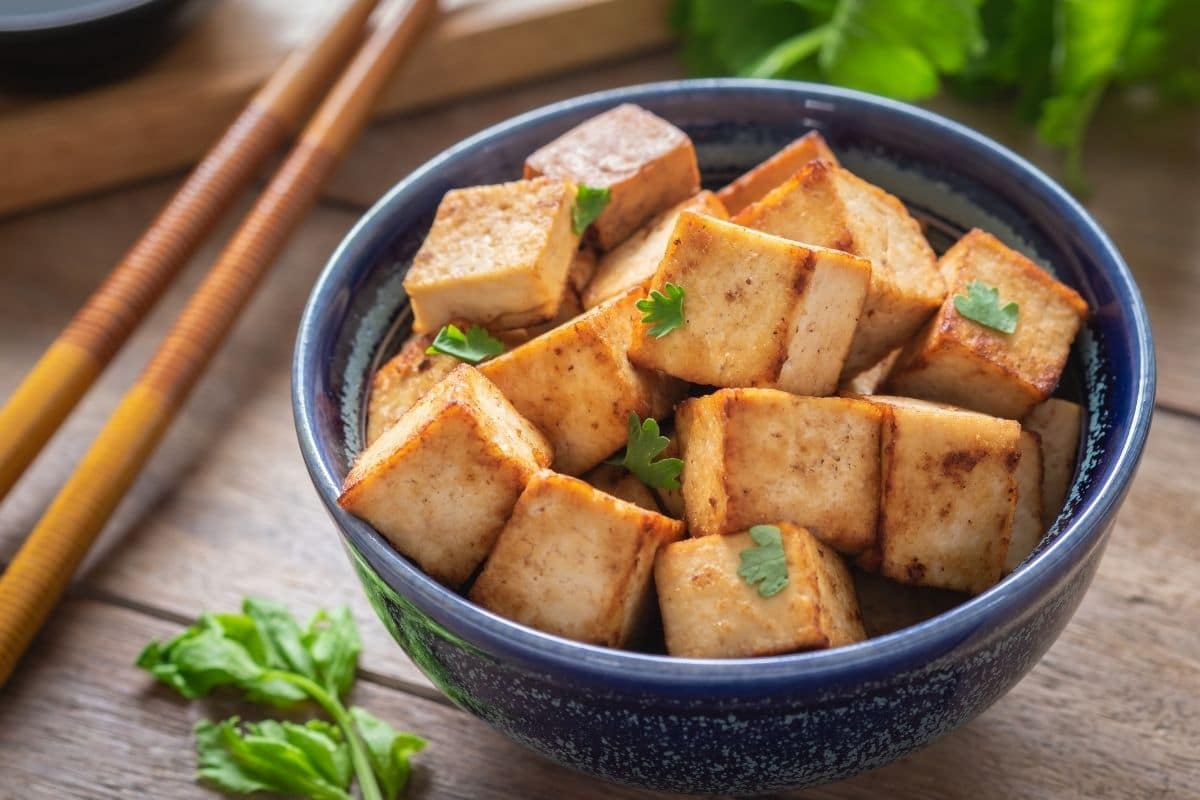Blue bowl of fried tofu on table with herbs and sticks.