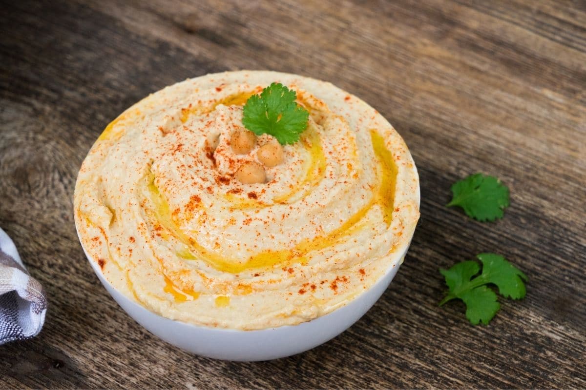 White bowl of hummus with herb on the top on wooden table.