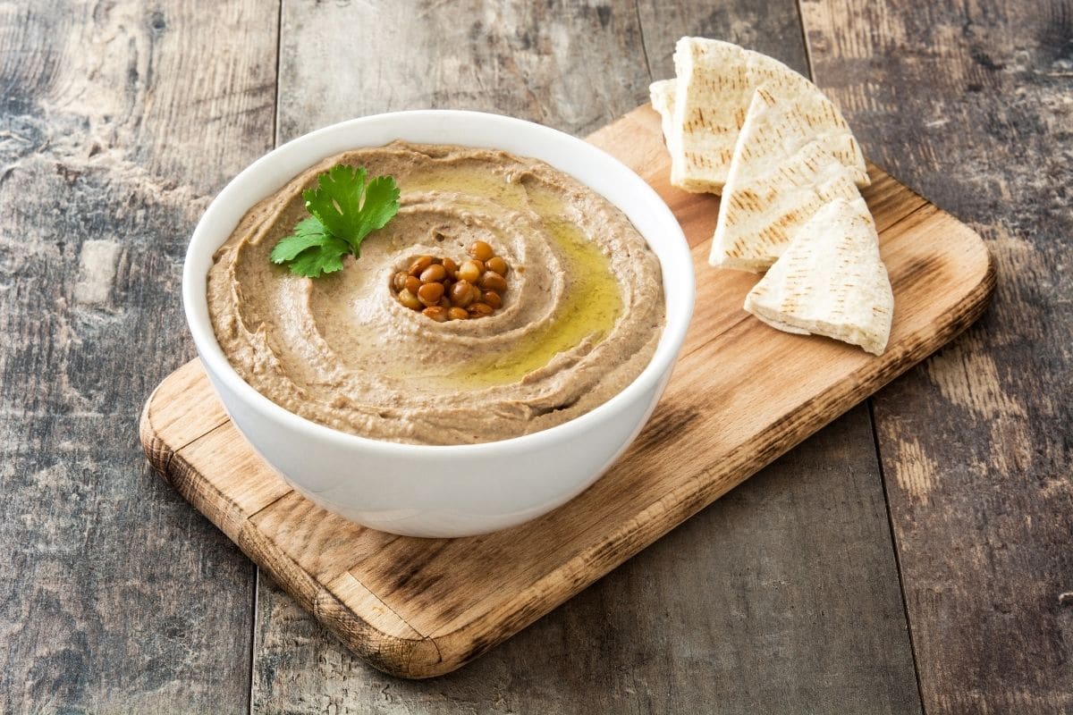 White bowl of hummus with slices of bread on wooden cutting board.