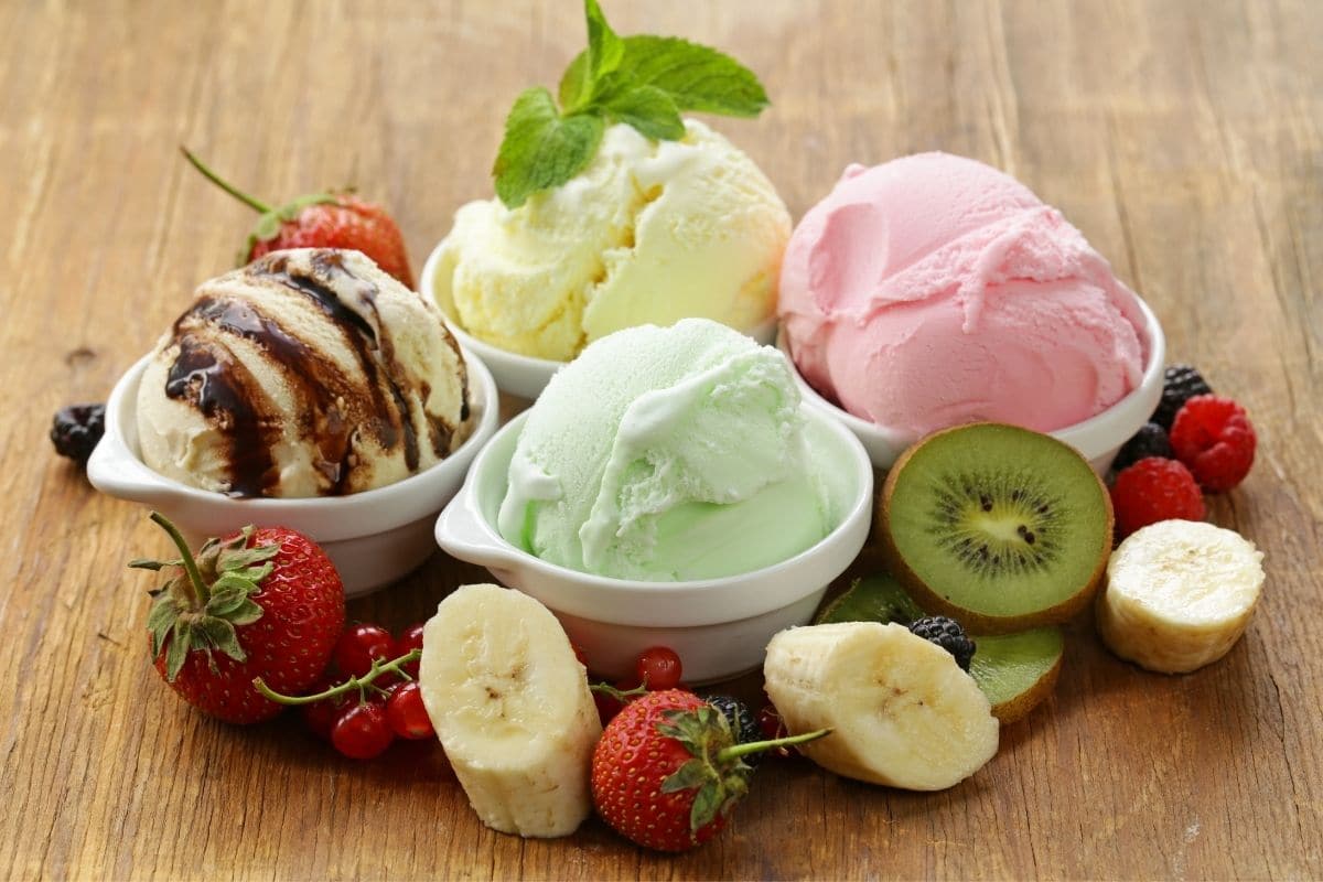 Small bowls of different flavors of ice cream with  sliced fruits on brown table.