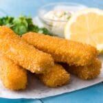 Fish sticks with herb, lemon and bowl of dip on white paper sheet on blue table.