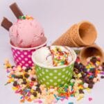 Paper cups full of ice cream with cones, sprinkles and chocolate chips scattered around.