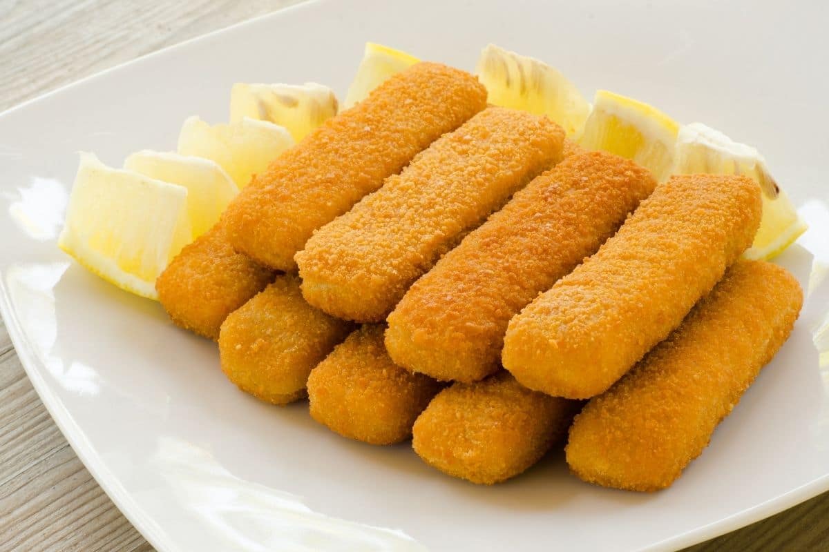 Fish sticks on white plate with pieces of lemon.