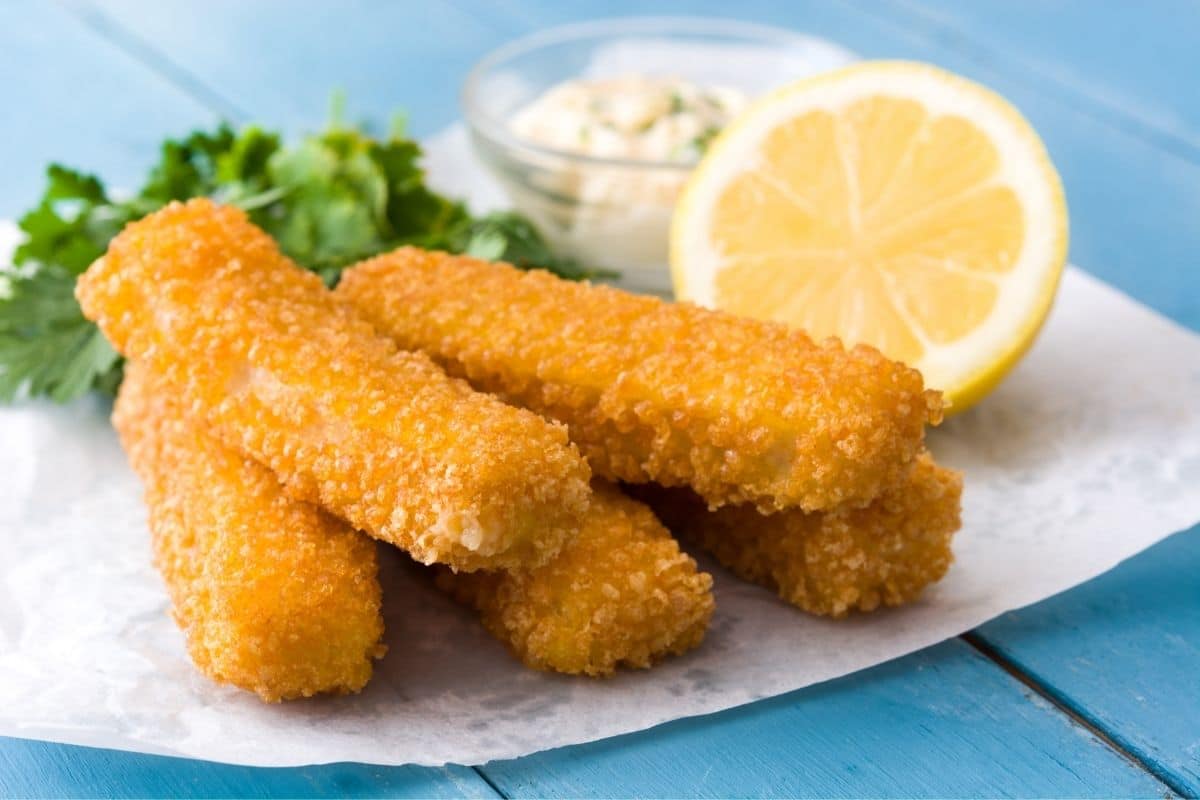 Fish sticks on white paper sheet with lemon, herb and dip on blue table.