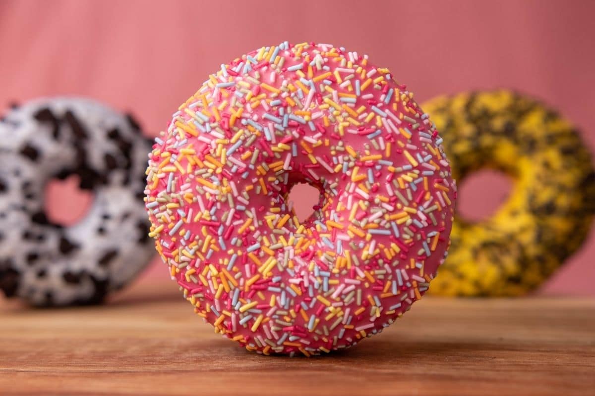 Pink sprinkled donut on wooden table, two donuts in the background.