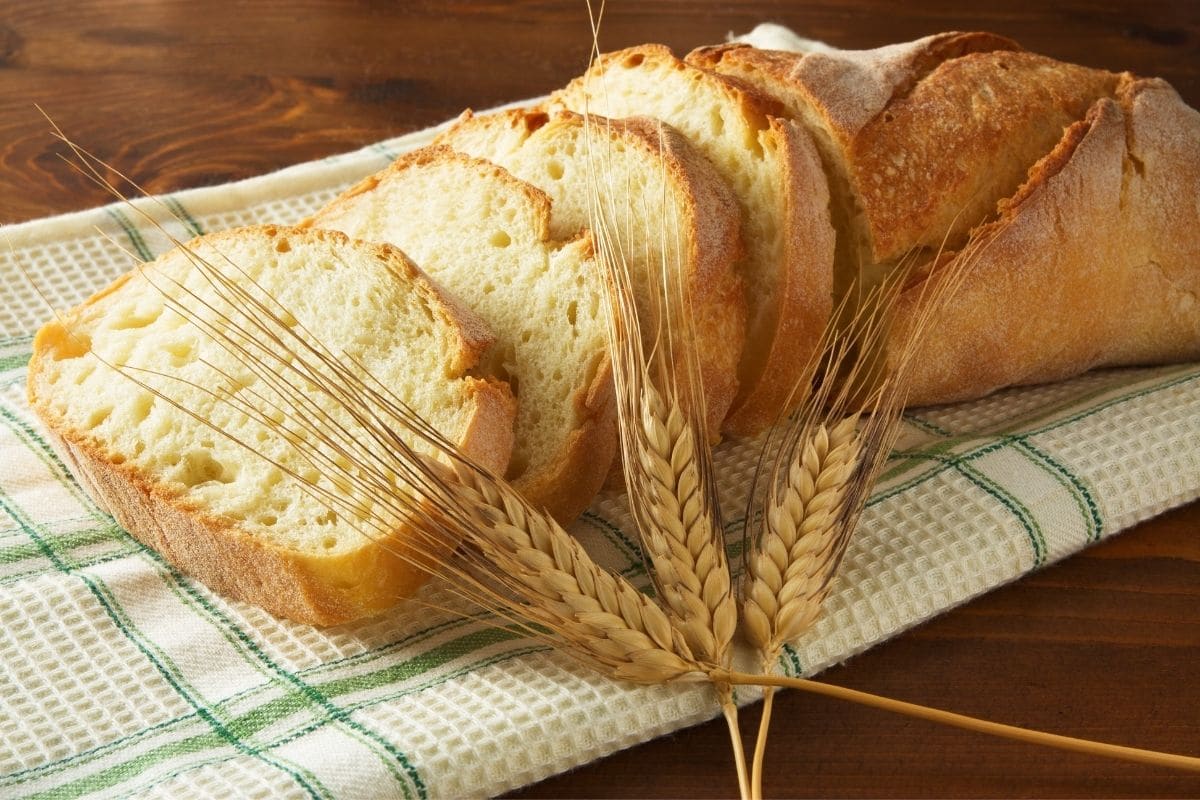 Partialy sliced loaf of bread on cloth wipe with grain stalk on wooden table.