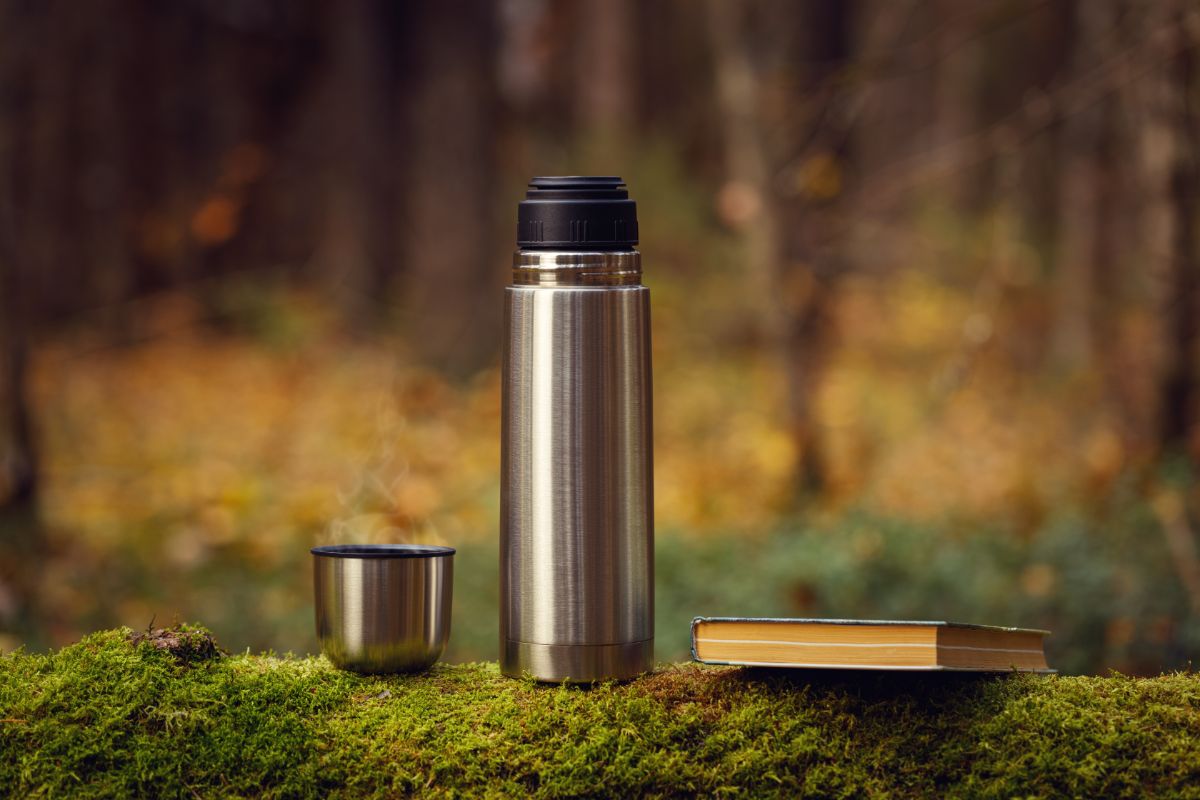 Stainless steel thermo bottle on wooden log with book and lid.