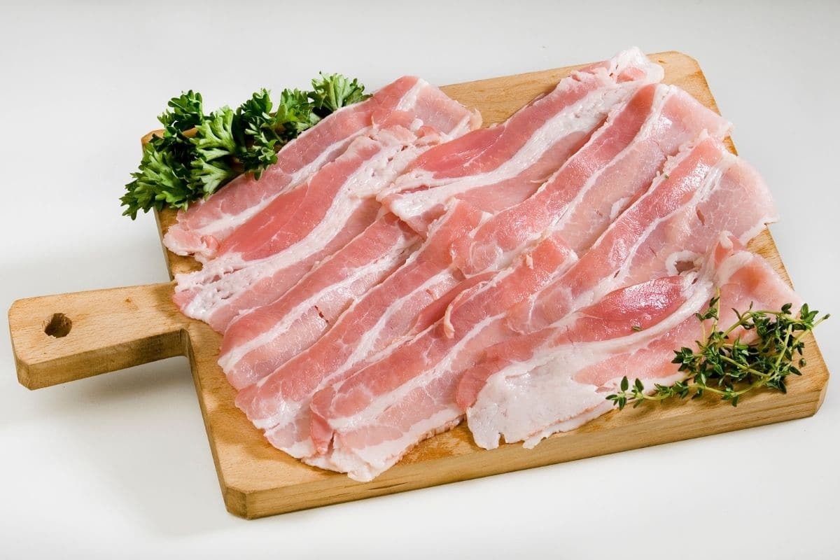 Stripes of bacon on wooden cutting board with herbs