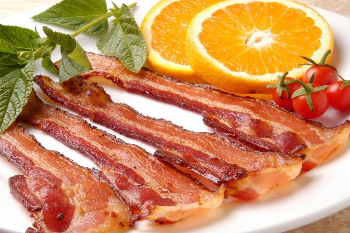 Crispy bacon stripe on white plate with tomatoes, slices of orange and herbs