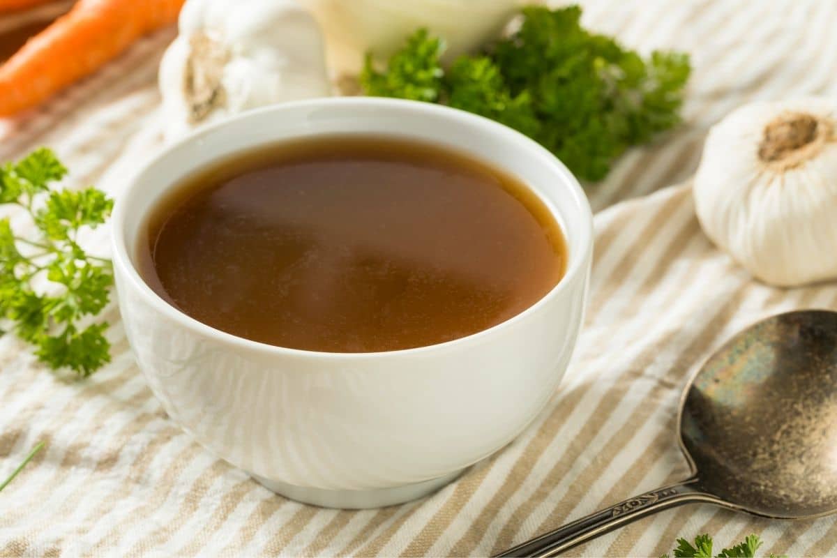 White bowl of bone broth on table with vegetable and spoon