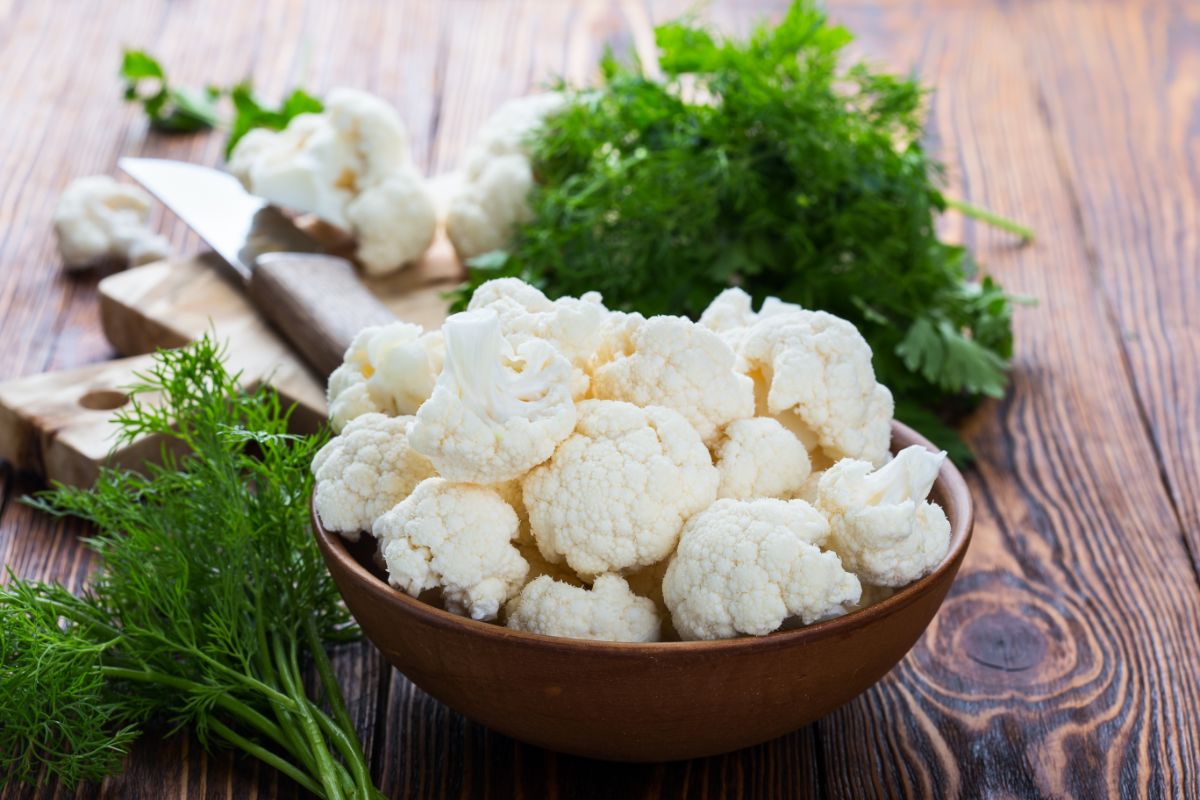 Wooden bowl of cauliflower  on wooden table with knife, herbs and cutting board