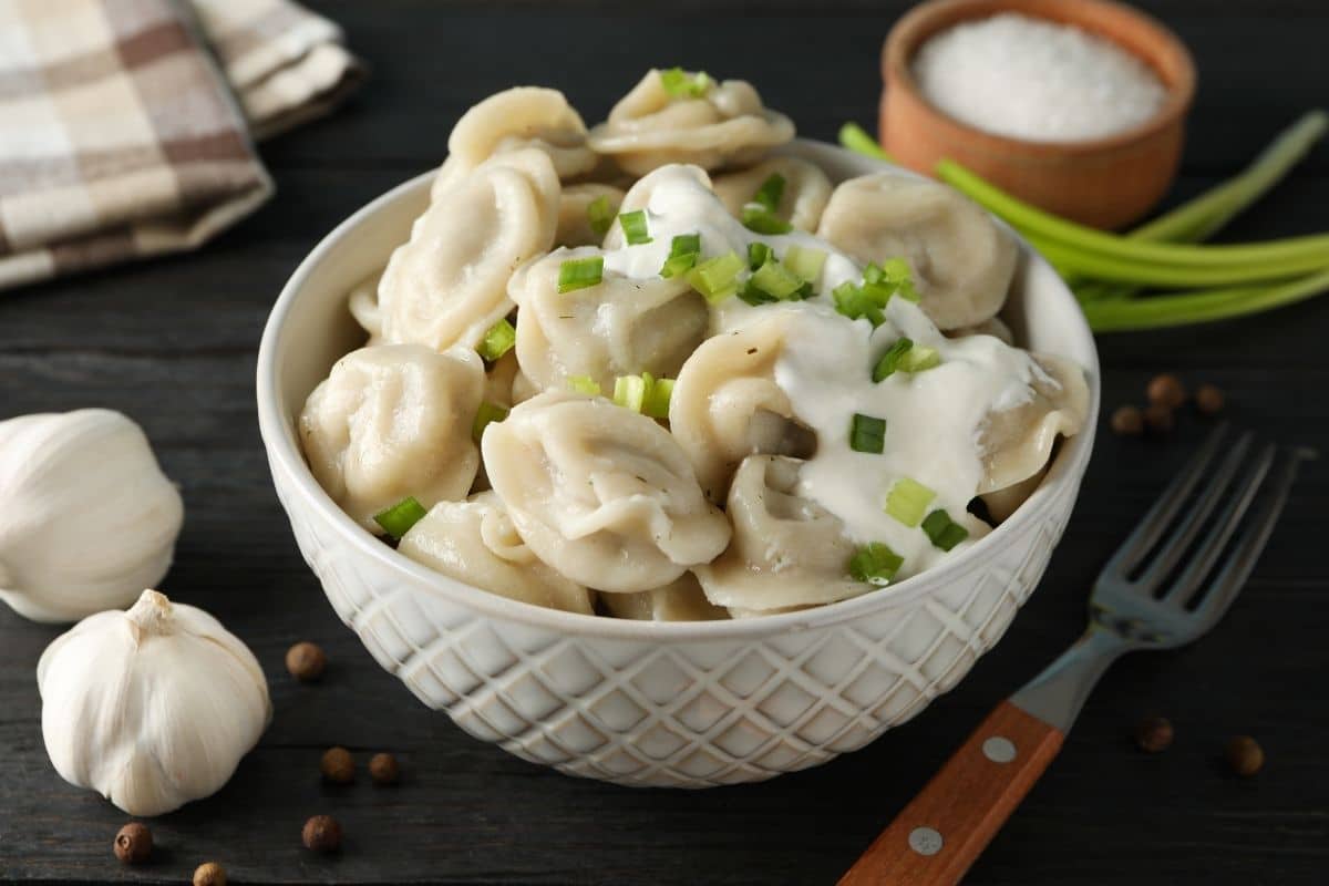 White bowl of fresh dumplings on table with fork and ingredients around.