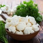 Wooden bowl of cauliflower on wooden table with herbs, cutting board and knife.