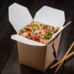 To-go box full of asian food on black table with wooden sticks