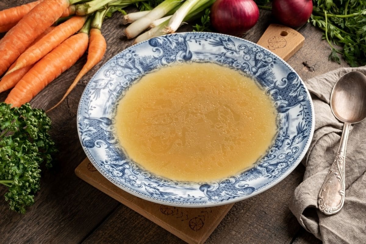 Blue-white plate of bone broth on table with vegetable and spoon around