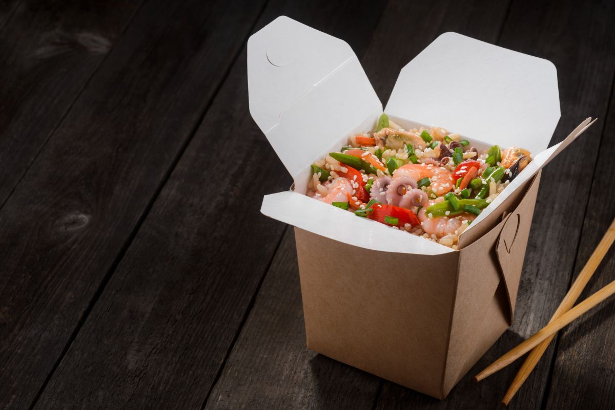 To-go box full of asian meal on wooden table with wooden sticks