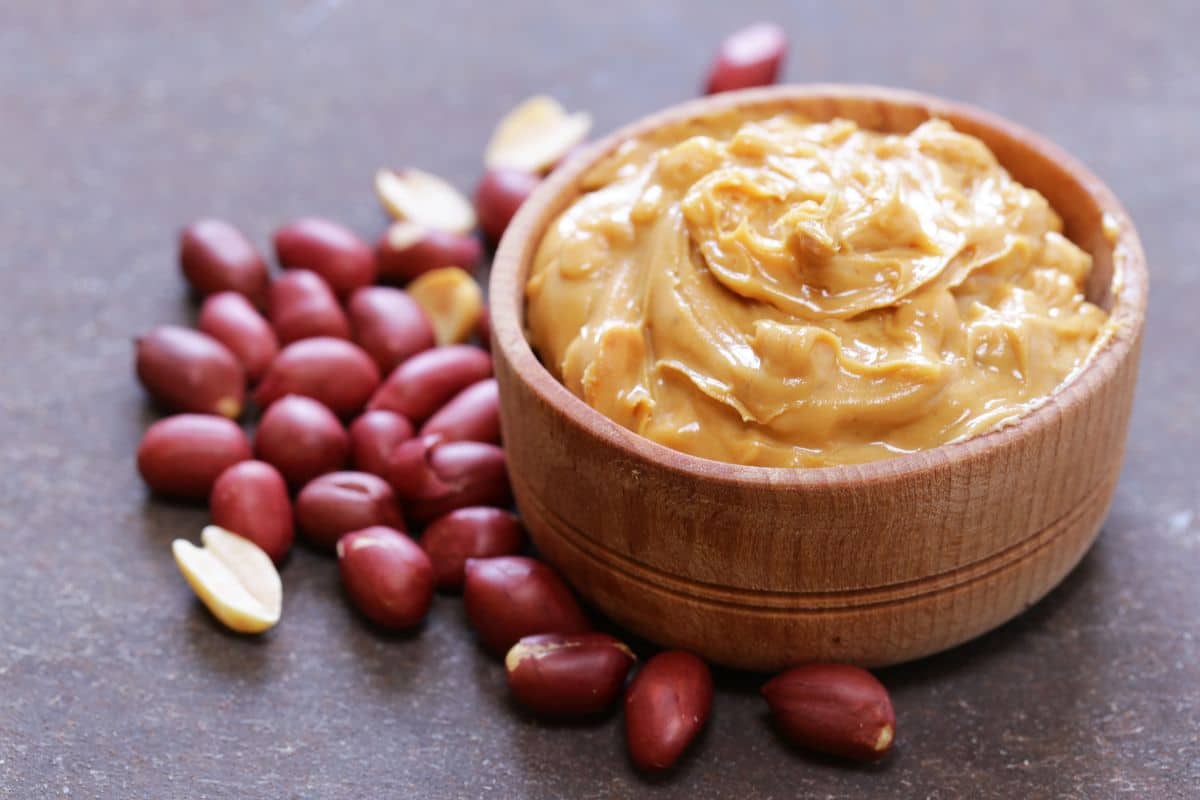 Wooden bowl of peanut butter with scattered peanuts on table