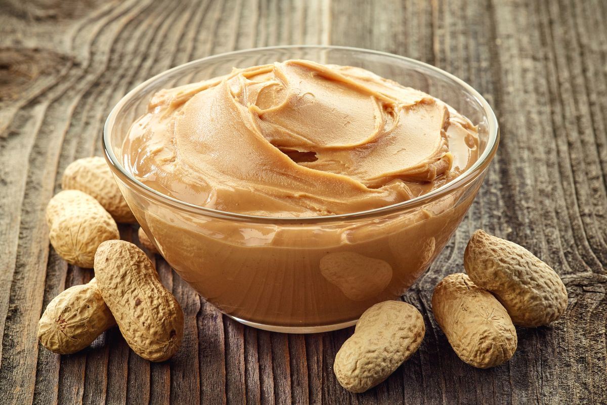 Glass bowl of peanut butter with scattered peanuts on wooden table