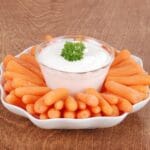 Glass bowl of ranch dressing surrounded by baby carrots on white plate