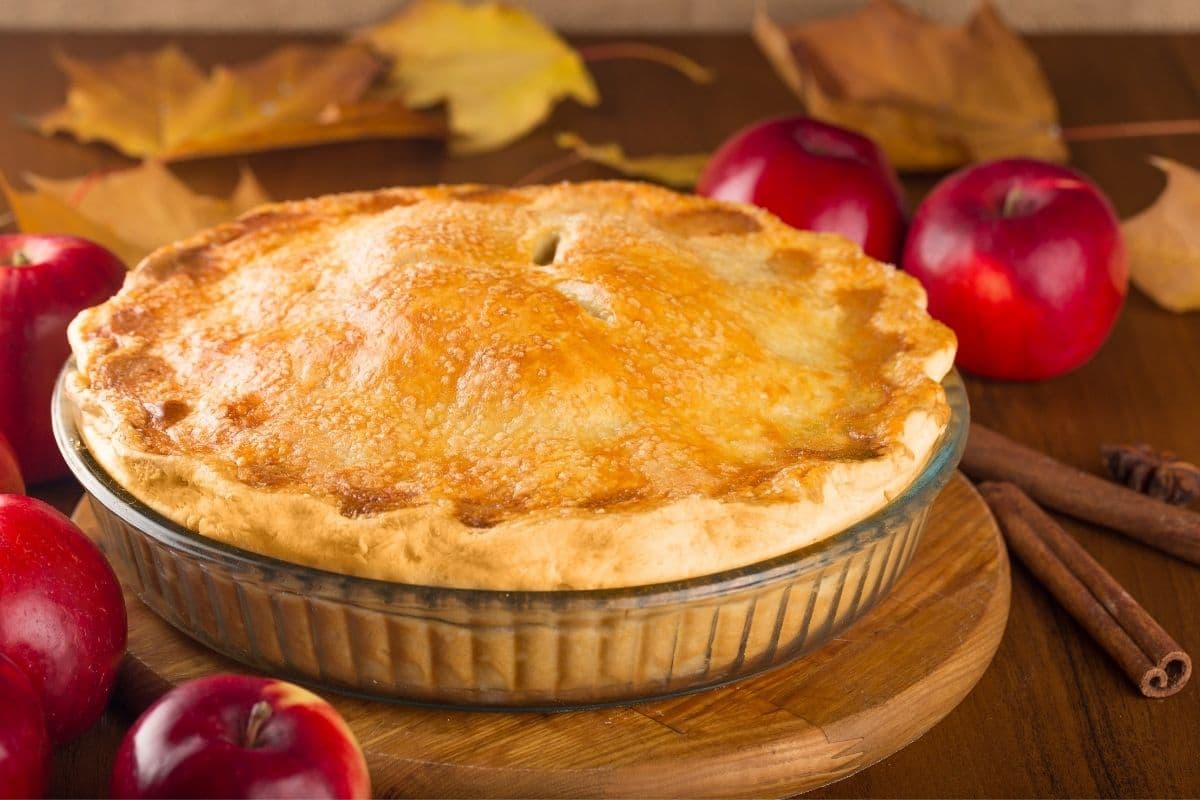 Apple pie on tray on wooden board on table with red apples, cinnamon sticks and leaves