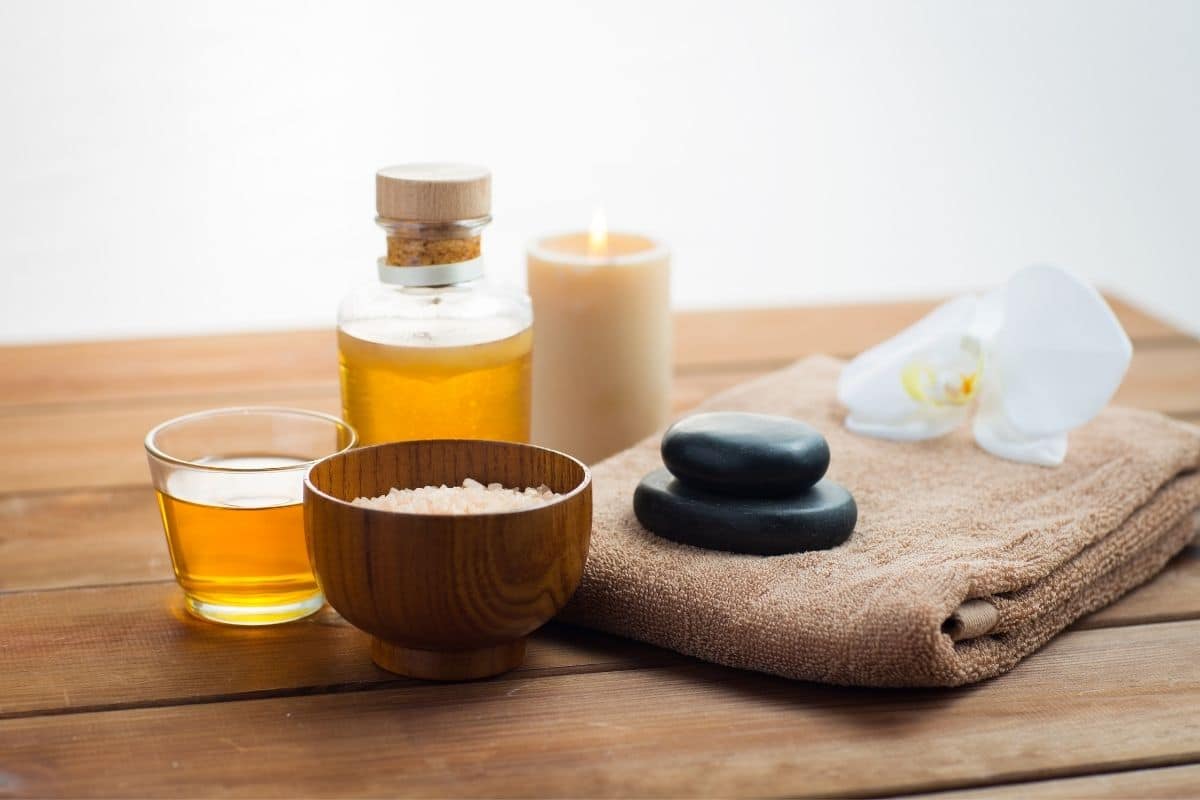 Glass jar and glass cup of massage oil with candy, bowl of salt, towel, flower on wooden table