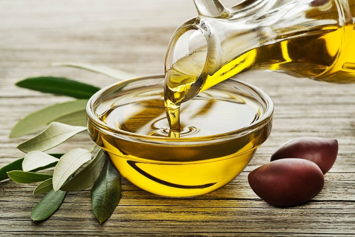 Olive oil poured from glass pitcher into glass bowl on wooden table