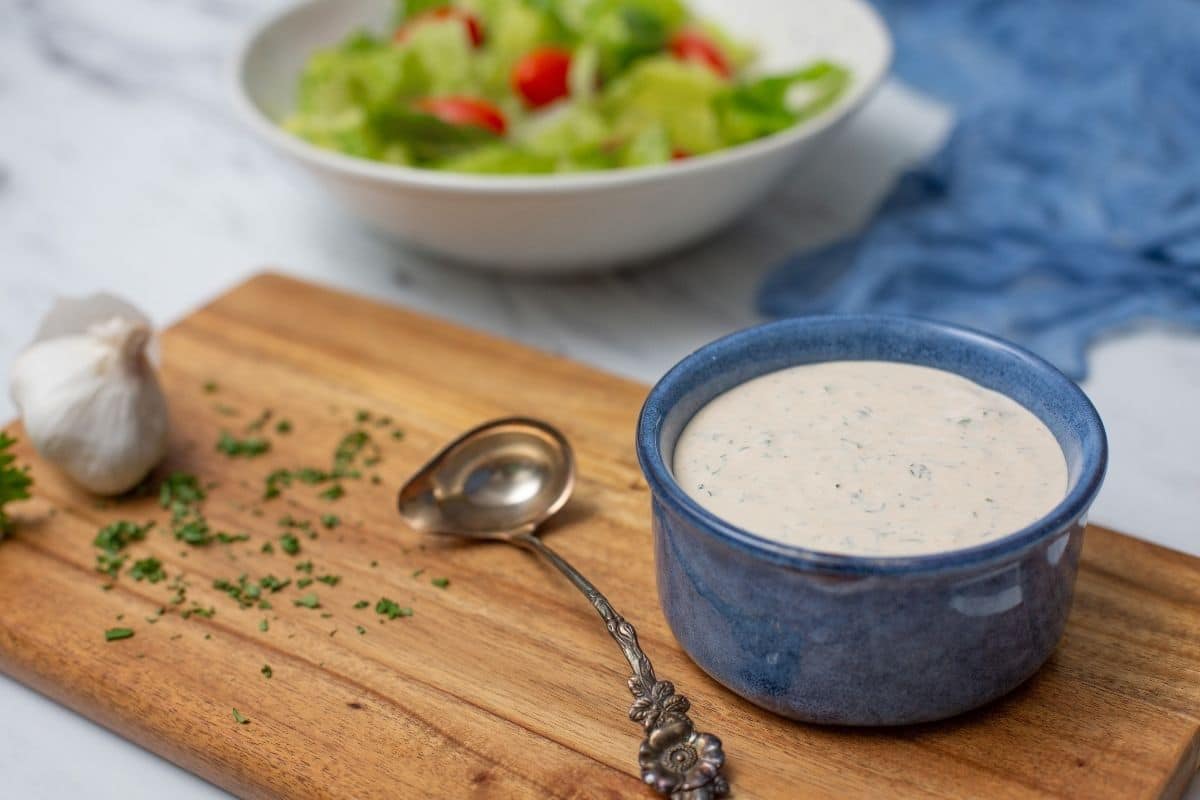 Ranch dressing in blue bowl on wooden cutting board with spoon and garlic