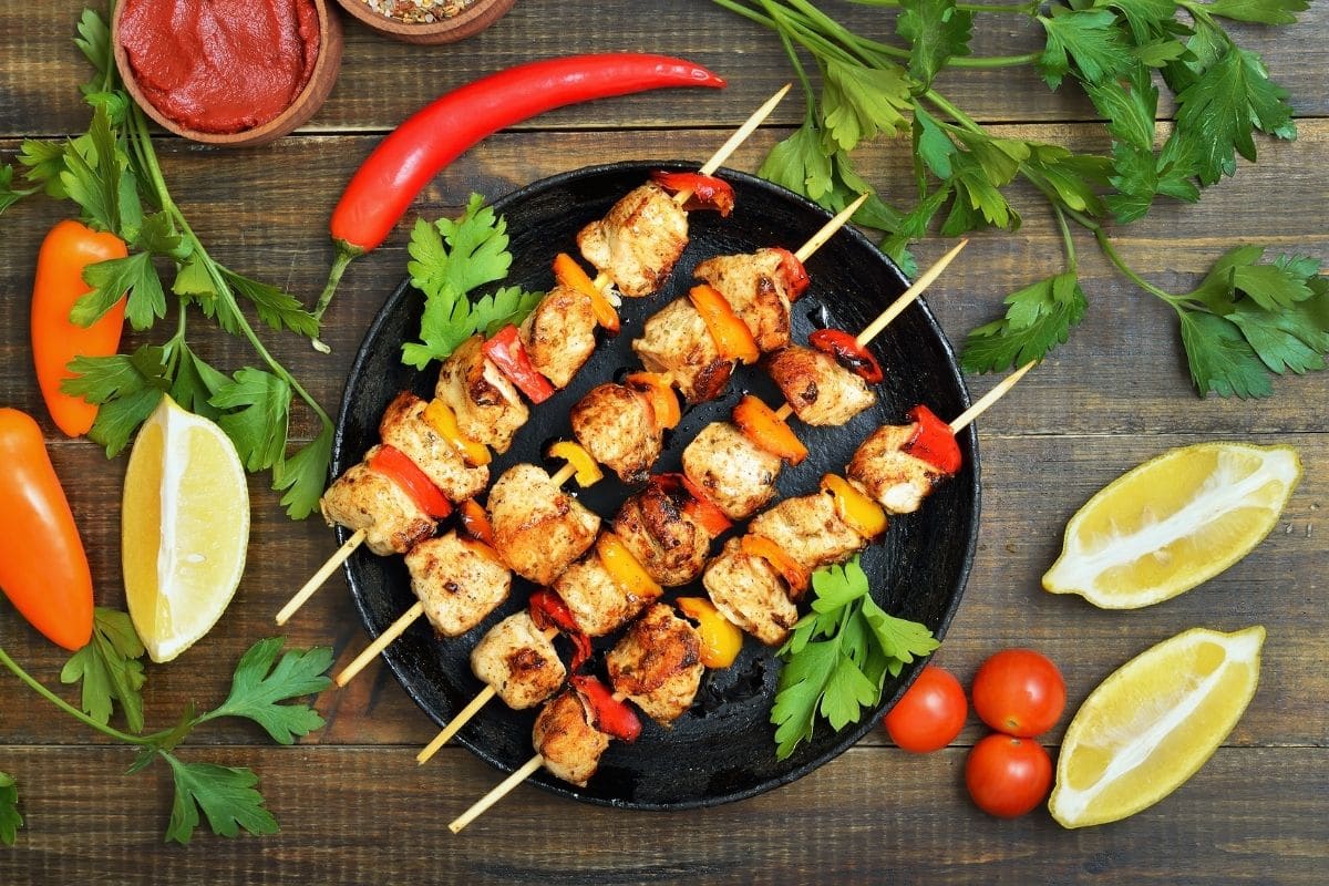 Kebab sticks on black pan with vegetables, lemon, and herbs on wooden table