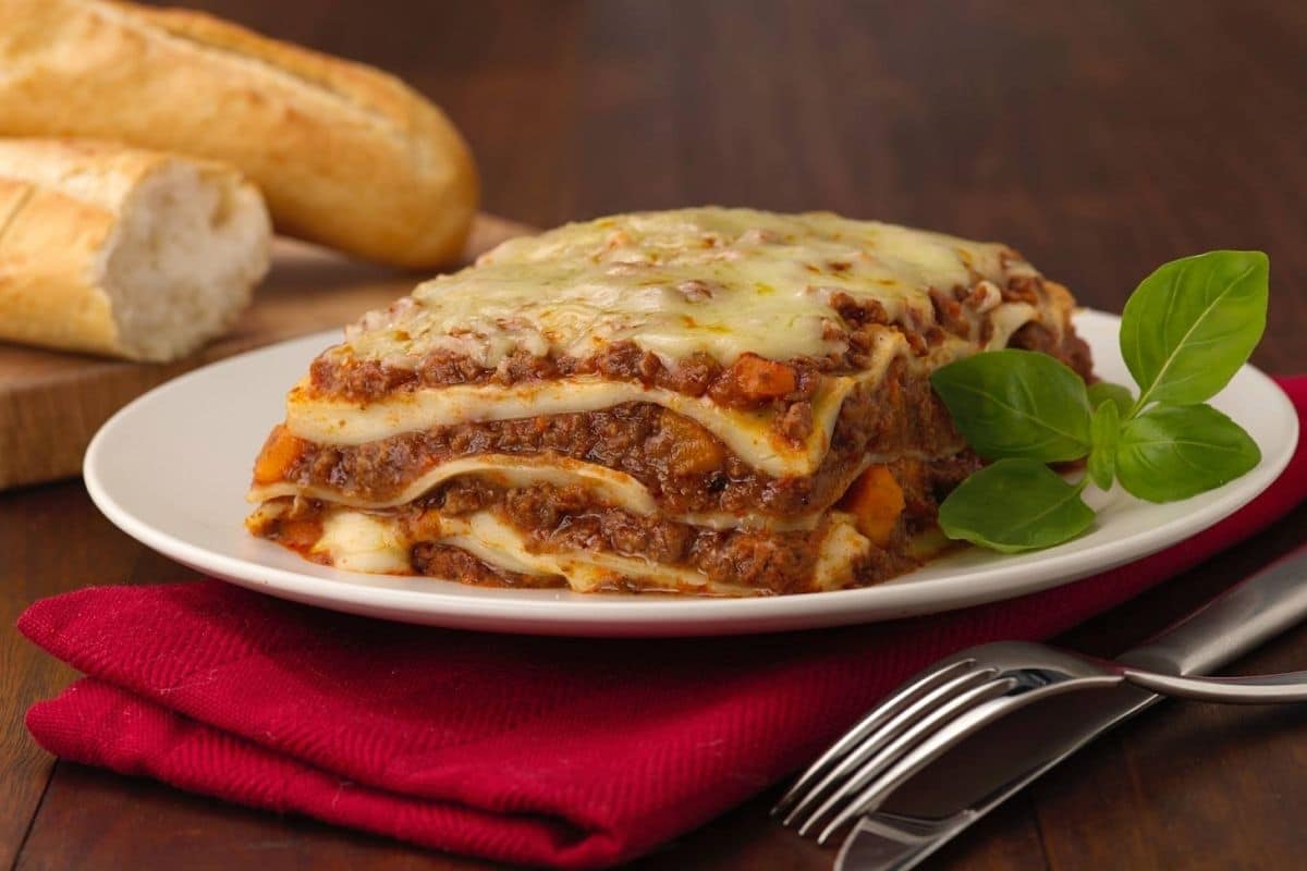 Lasagna on white plate with herb on red cloth wipe, knife and fork on brown table