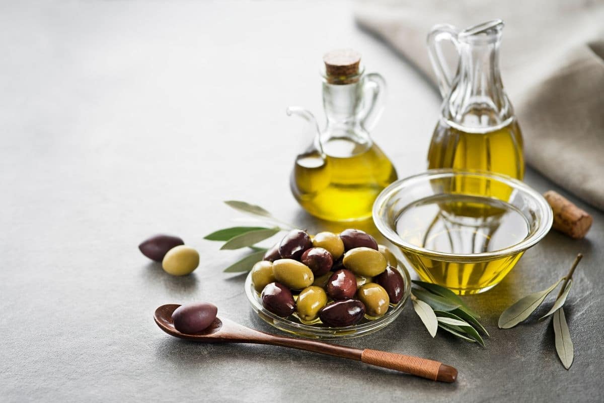 Glass pitchers of olive oil,glass bowl of olive oil and bowl of olives and wooden spoon on gray table