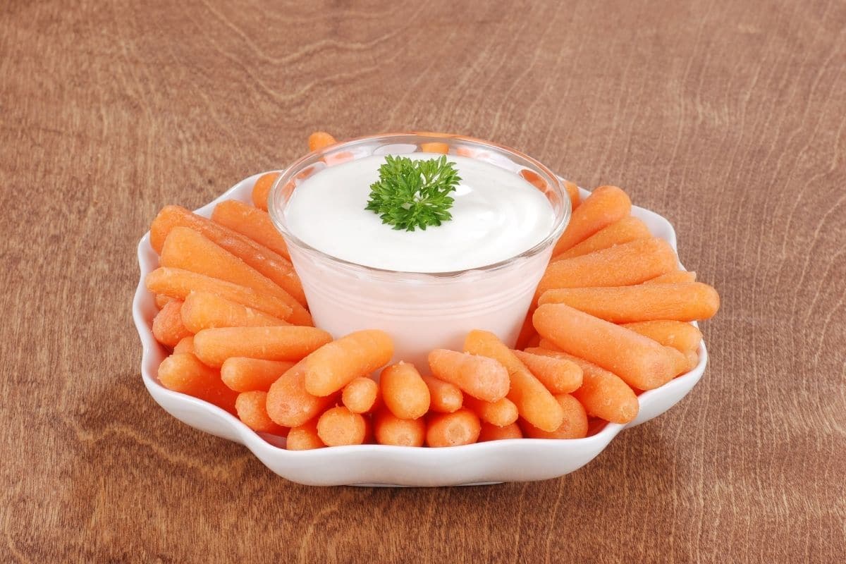 Ranch dressing in glass bowl surrounded by baby carrots on white plate