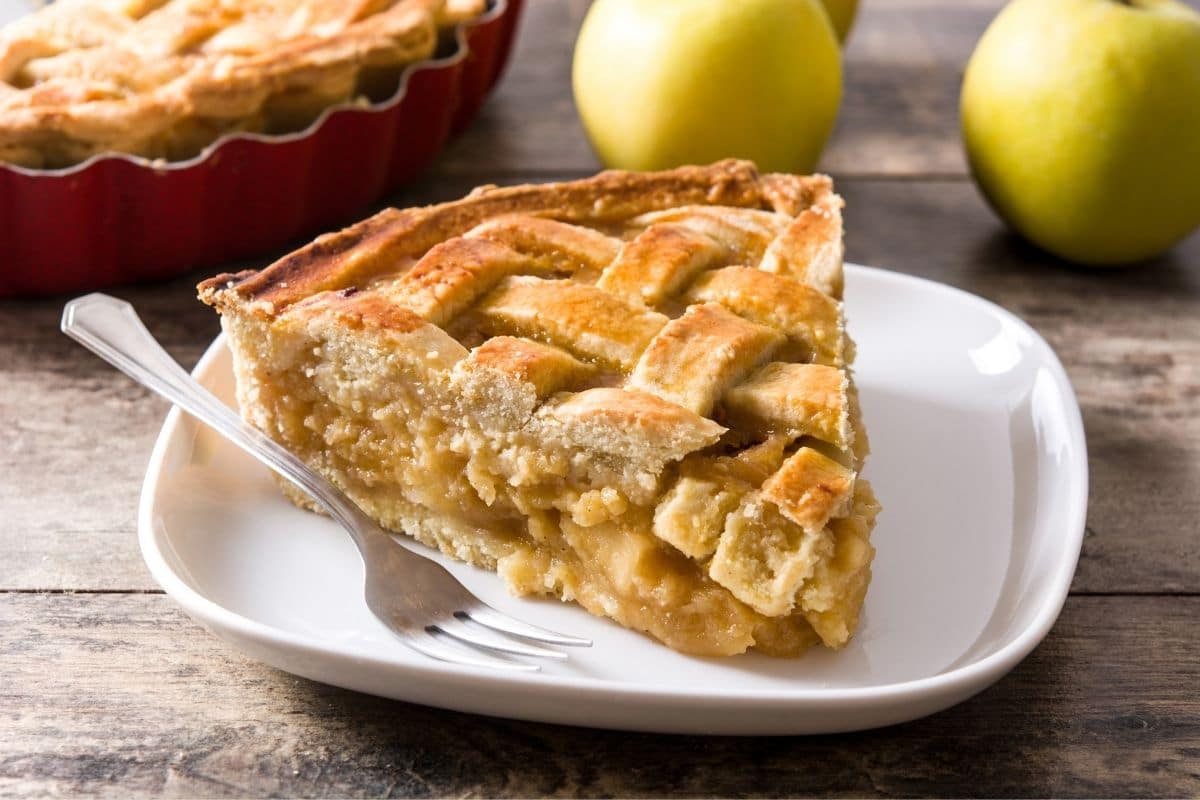 Slice of apple pie with fork on white plate, rest of pie and apples in the background