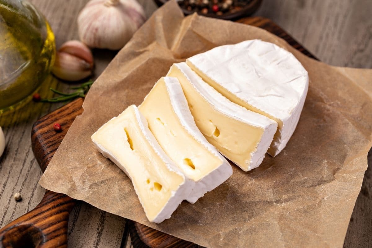 Sliced brie cheese on paper sheet on wooden cutting board with ingredients around