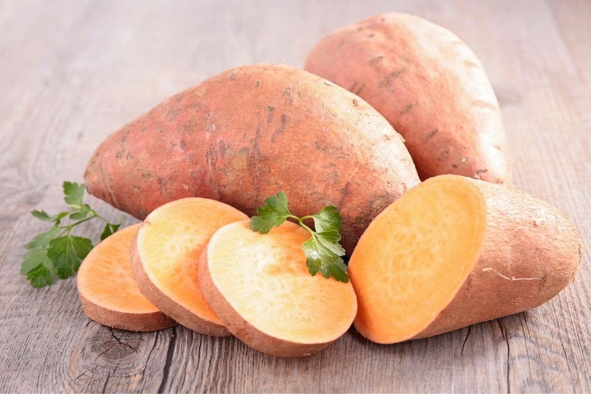 Two whole swwet potatoes and one sliced on wooden table with herbs