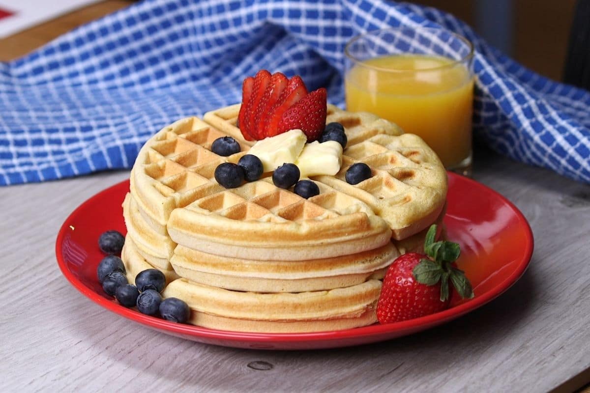 Waffles with butter, fruits on red plate  on gray table with glass cup of orange juice and cloth wipe