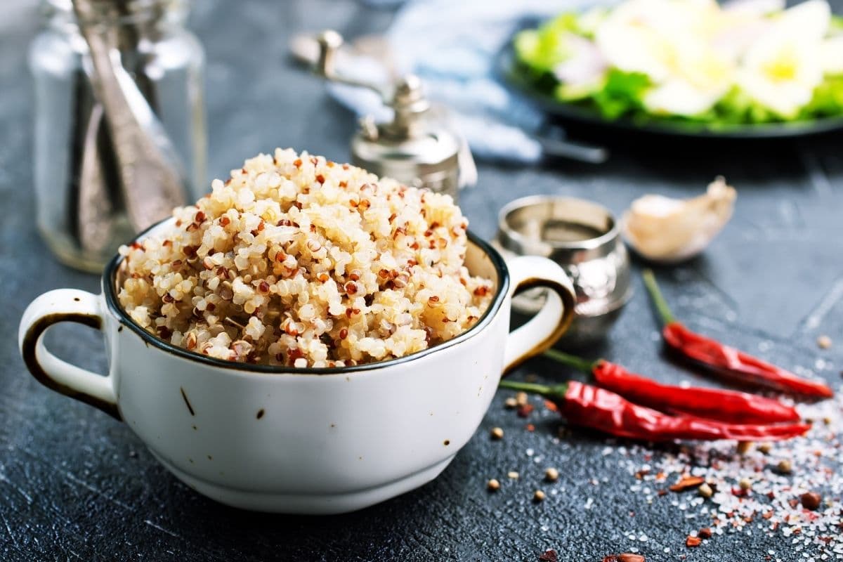 White pot of cooked quinoa on table with chilli peppers, salt, and other ingredients on black table