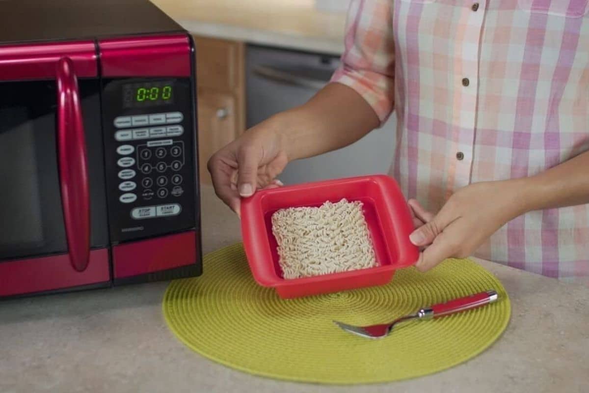 Man holding red microwave pasta cooker nexto to mcirowave over table with fork