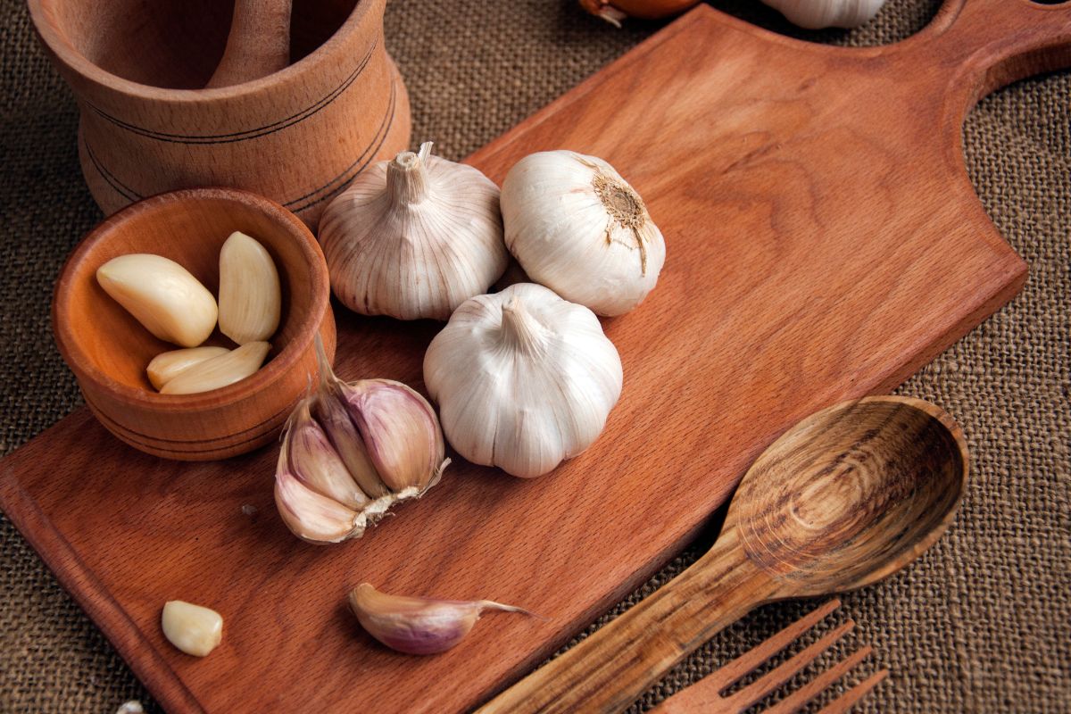 Trimmed garlic in a woode bowl next to garlic heads on  a board and wooden spoon, fork and cup.